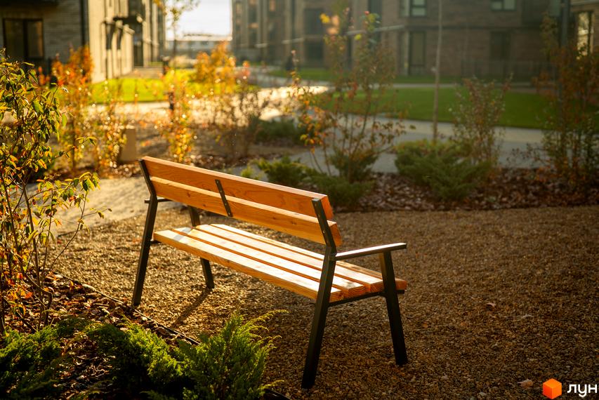 Зображення лавки на території Житлового комплексу Долішній. На задньому фоні видно будівлі та зелені насадження.