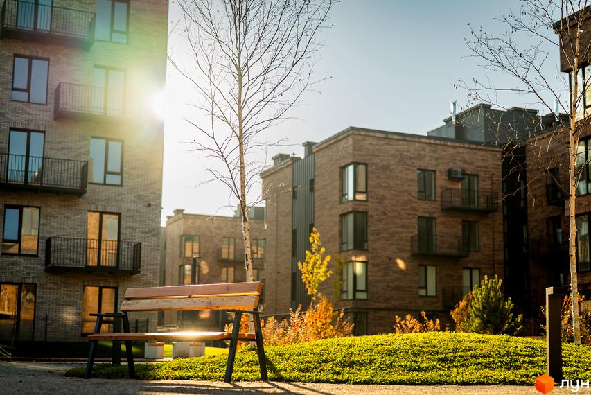 Зображення фасаду Житлового комплексу Долішній та лавочки на прибудинковій території. Видно дерева та газон.