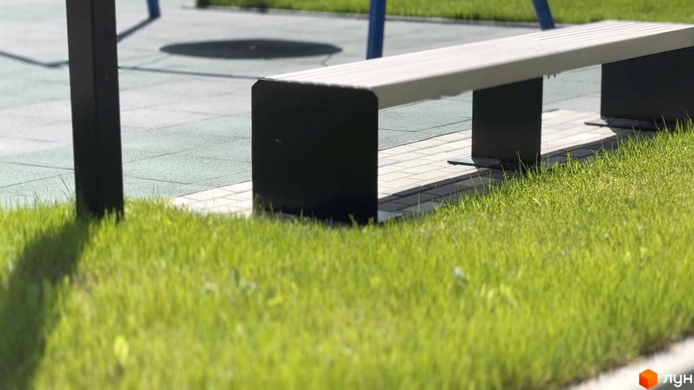 Фото лавки біля Житлового комплексу Стандарт. Навколо зелений газон, а позаду видно дитячий майданчик.
