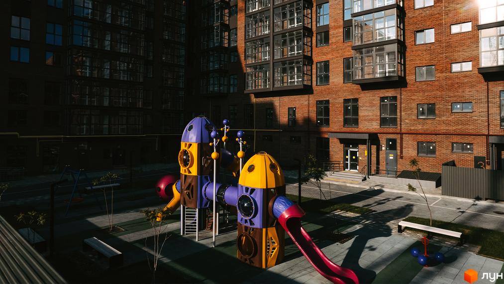 Вигляд на дитячий майданчик та цегляний будинок у Житловому комплексі Стандарт. На території є лавки та дерева.