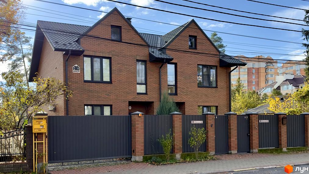 Зовнішній вигляд дуплексу в Котеджному містечку PARK PLACE з червоної цегли та чорною покрівлею. Перед будинком сіра огорожа та зелені насадження.