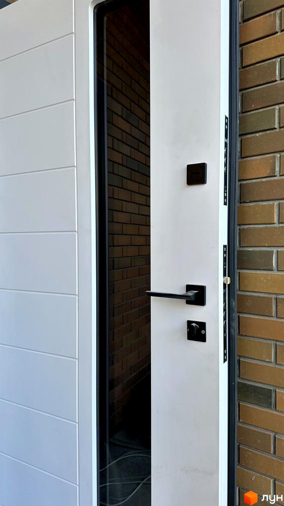 Зображення вхідних дверей у дуплекс PARK PLACE. Білі двері з чорною фурнітурою на фоні цегляної стіни.
