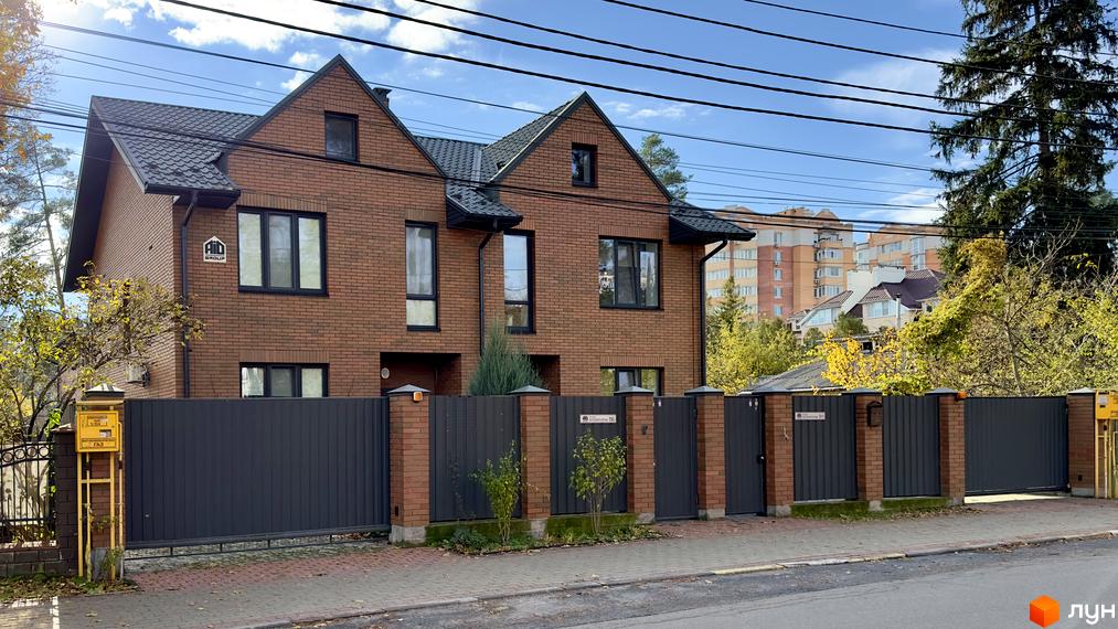 Зображення фасаду двоповерхового будинку з червоної цегли в Котеджному містечку PARK PLACE. Перед будинком сірий паркан та зелені насадження.