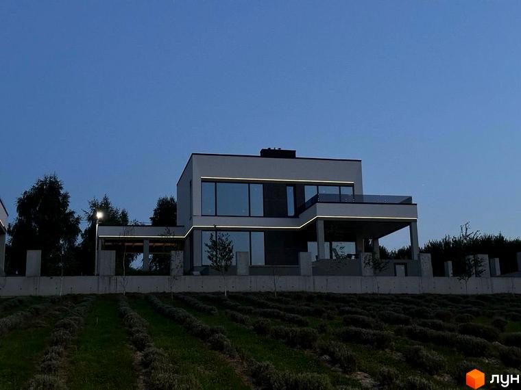 Двоповерховий будинок з великими вікнами та підсвіткою на фасаді. Перед будинком розташована доглянута ділянка з рослинами.