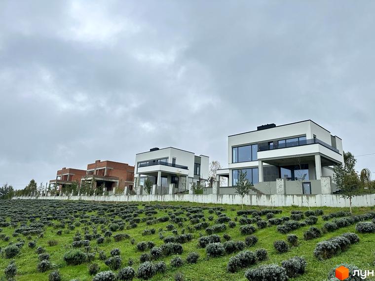 Білі будинки з панорамними вікнами в Містечко котеджів Belvedere на тлі хмарного неба. На передньому плані газон з кущами.