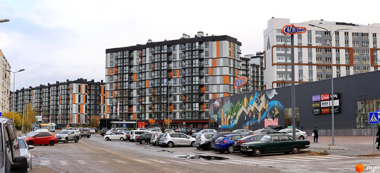 Багатоповерховий будинок ЖК Атмосфера з фасадом в сірих, білих та помаранчевих кольорах. Перед будинком парковка з автомобілями.