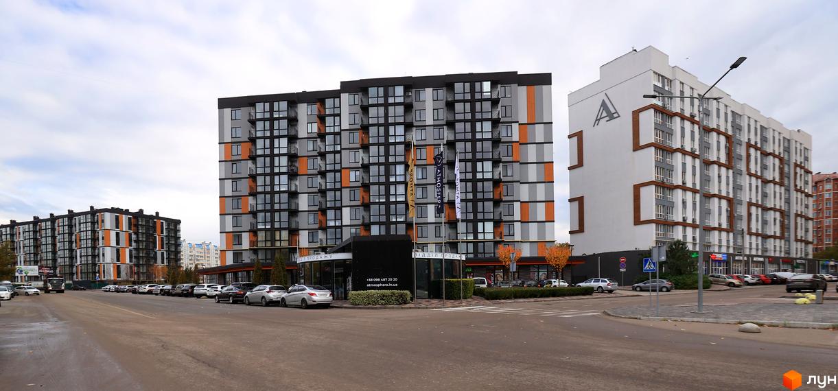 Багатоповерховий будинок ЖК Атмосфера з фасадом у сіро-чорних тонах з помаранчевими вставками. На прибудинковій території є парковка та зелені насадження.