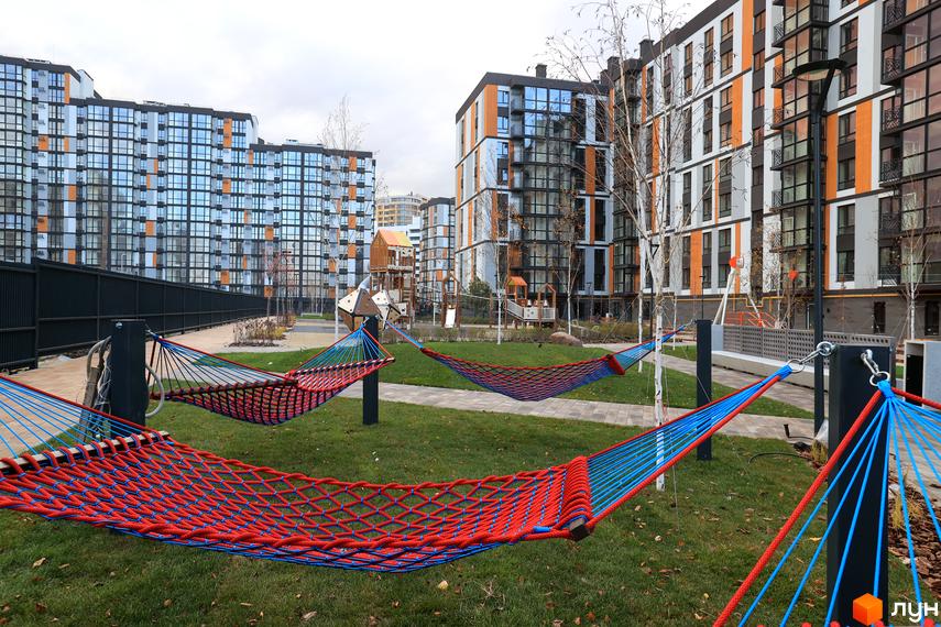 Житловий комплекс ATMOSFERA LIFE з фасадами в помаранчевих та сірих тонах. На території комплексу розташовані гамаки для відпочинку та дитячий майданчик.