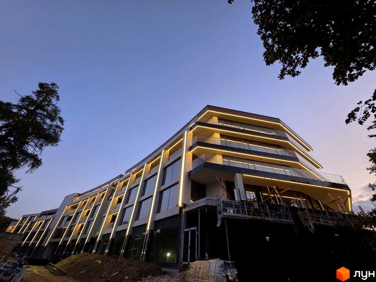 Будівельний проєкт Emily Apartments з підсвіткою фасаду та скляними балконами на тлі вечірнього неба. Навколо новобудови видно будівельні роботи та дерева.