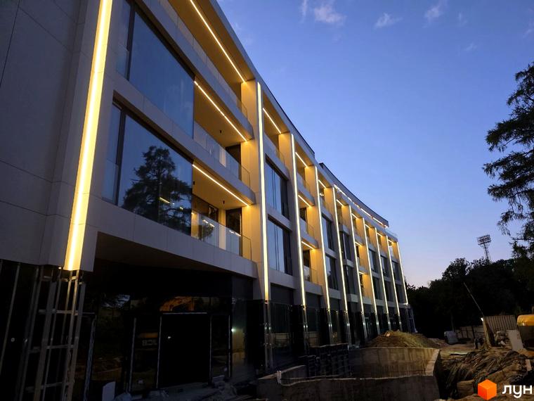 Житловий комплекс Emily Apartments з підсвіченим фасадом та скляними балконами на тлі вечірнього неба. На передньому плані видно будівельні роботи.