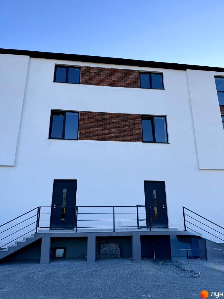 Новобудова ЖК Затишок з білими фасадами, цегляними вставками та темними вікнами. Перед будинком розташовані сходи з чорними перилами та бруківка.