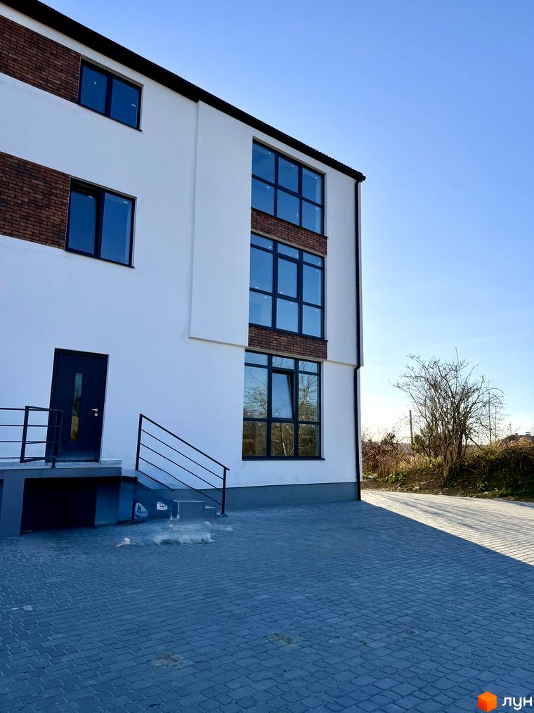 Зовнішній вигляд новобудови ЖК Затишок з білими стінами, цегляними вставками та великими вікнами. Поруч брукована прибудинкова територія та зелені насадження.