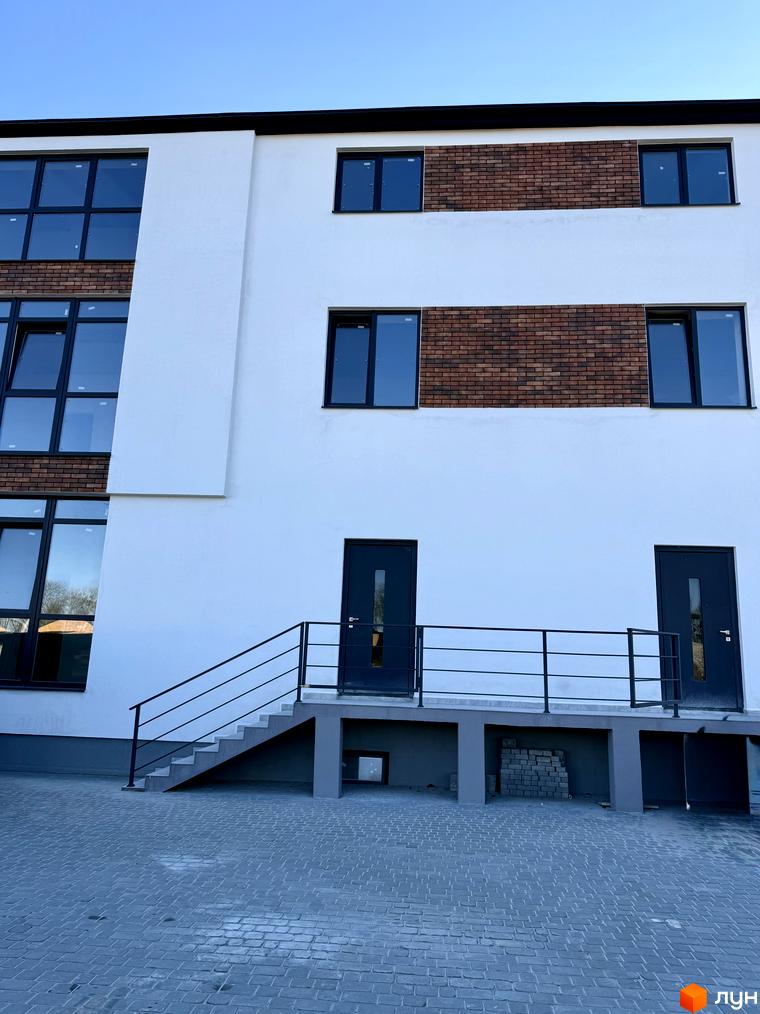 Зовнішній вигляд новобудови ЖК Затишок з білими стінами, цегляними вставками та темними вікнами. Поруч розташована брукована прибудинкова територія зі сходами та дверима.