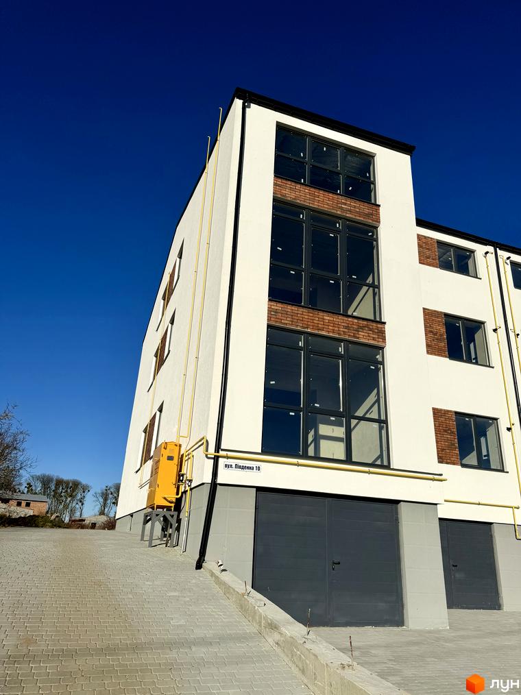 Зовнішній вигляд новобудови ЖК Затишок з цегляними вставками та великими вікнами. Прибудинкова територія з бруківки та гаражними воротами.