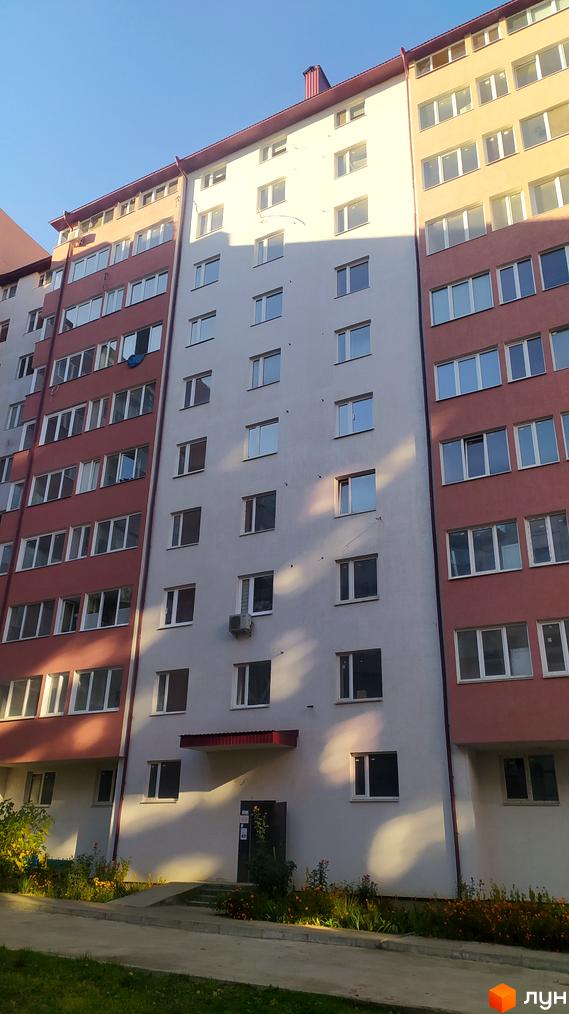 Багатоповерховий житловий комплекс з фасадом у червоних та білих тонах. Поруч розташовані зелені насадження та квіти.