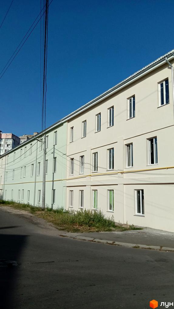 Зовнішній вигляд житлового комплексу на Будівельному: двоповерхові будинки світло-бежевого та світло-зеленого кольору з білими вікнами. Перед будинками розташована дорога та зелені насадження.