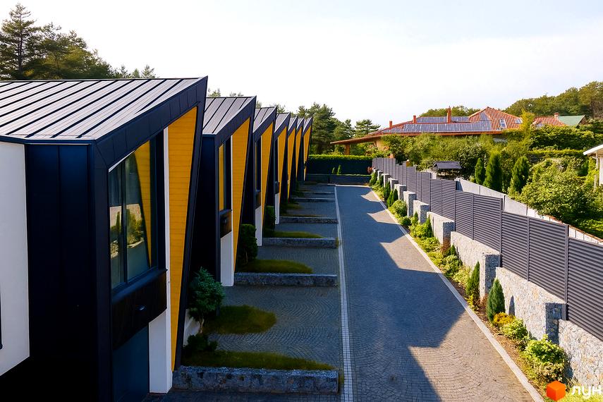 Таунхаус ДЖЕРЕЛЬНА: фасади будівель з жовтими акцентами та чорними дахами, асфальтована дорога та огороджена територія. Зовнішній вигляд новобудови та прибудинкової території.