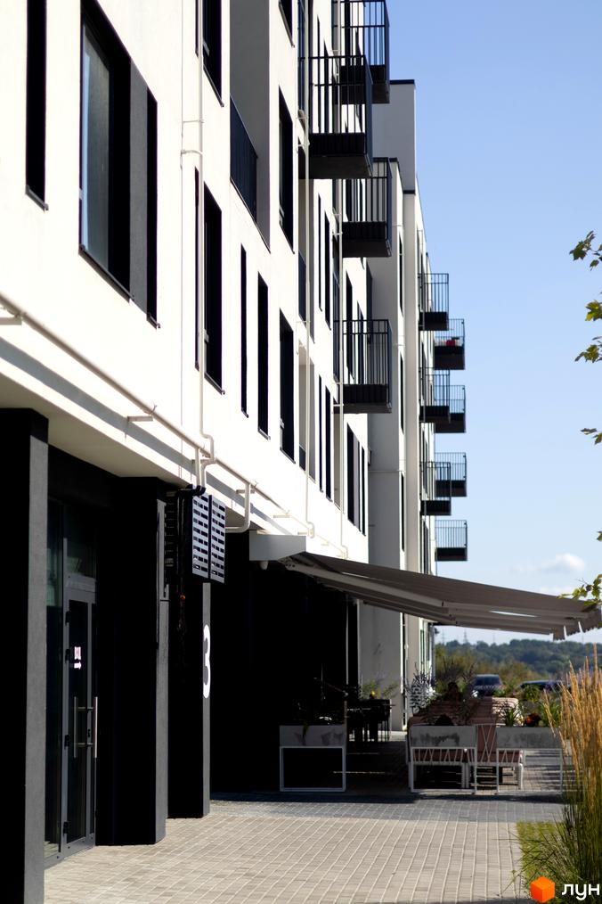 Житловий комплекс Empatika: біла фасадна стіна з чорними вікнами та балконами, прибудинкова територія з терасою та рослинами.