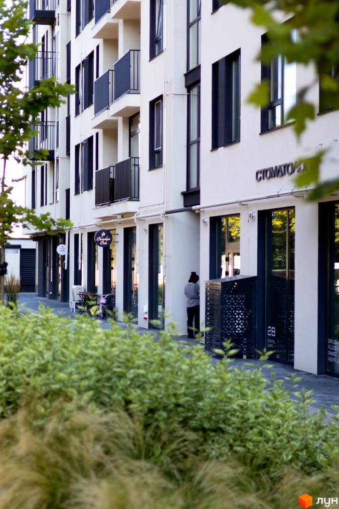 Зовнішній вигляд новобудови ЖК Empatika з фасадом білого кольору та темними вікнами. Поруч розташовані зелені насадження та тротуар.