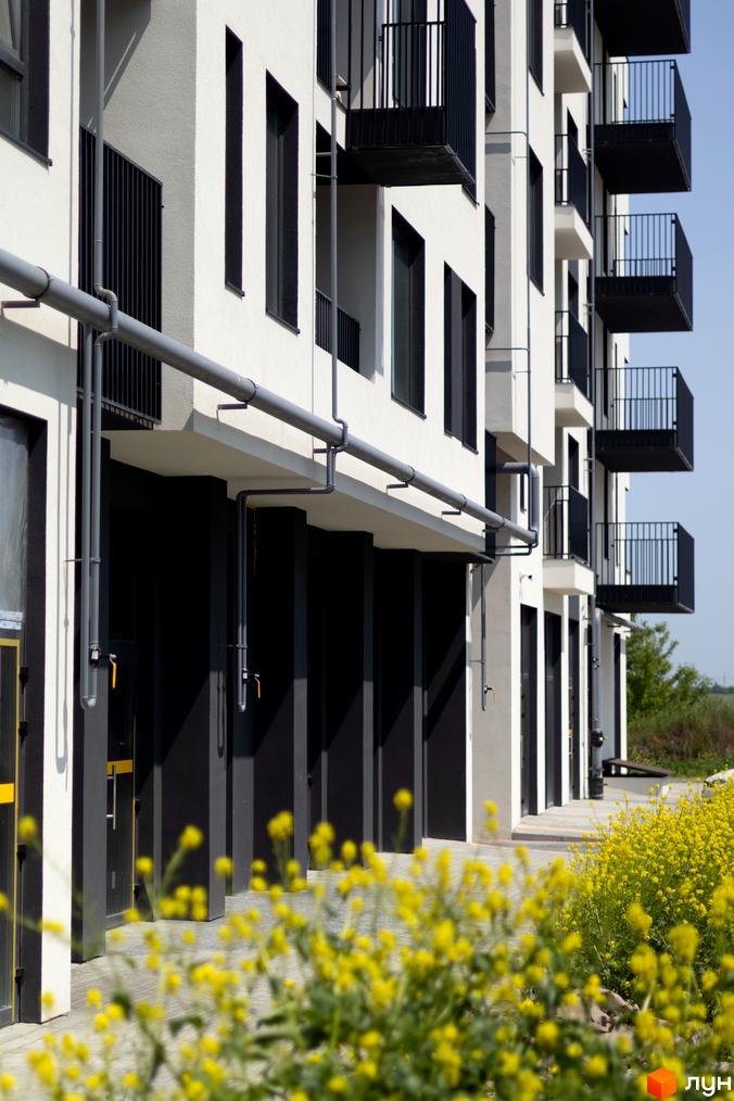 Зовнішній вигляд новобудови ЖК Empatika з балконами та трубами, поруч жовті квіти. Зовнішній вигляд будинку та прибудинкової території.