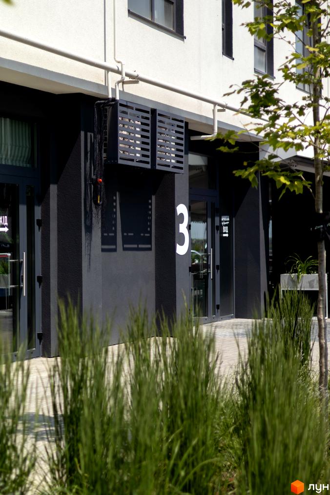 Зовнішній вигляд новобудови ЖК Empatika з білими та чорними фасадами, вікнами та дверима. Поруч розташована прибудинкова територія з деревами та травою.