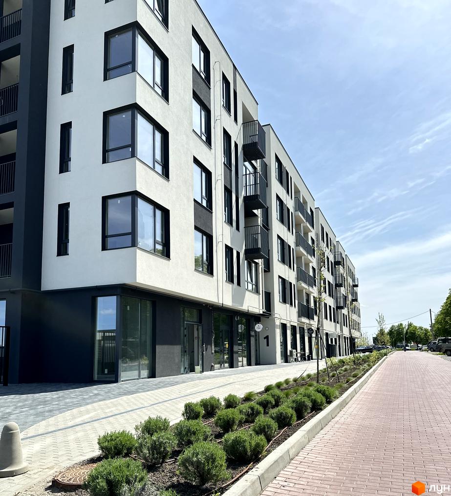 Зовнішній вигляд будинку ЖК Empatika з білими фасадами, чорними вікнами та балконами. Прибудинкова територія з бруківкою та зеленими насадженнями.