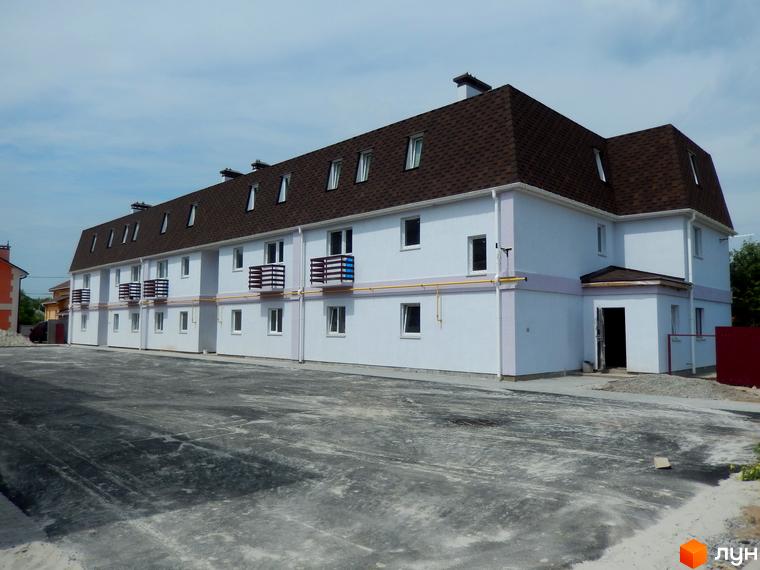 Зображення житлового комплексу. Двоповерховий будинок з балконами та мансардним дахом. Перед будинком асфальтований майданчик.