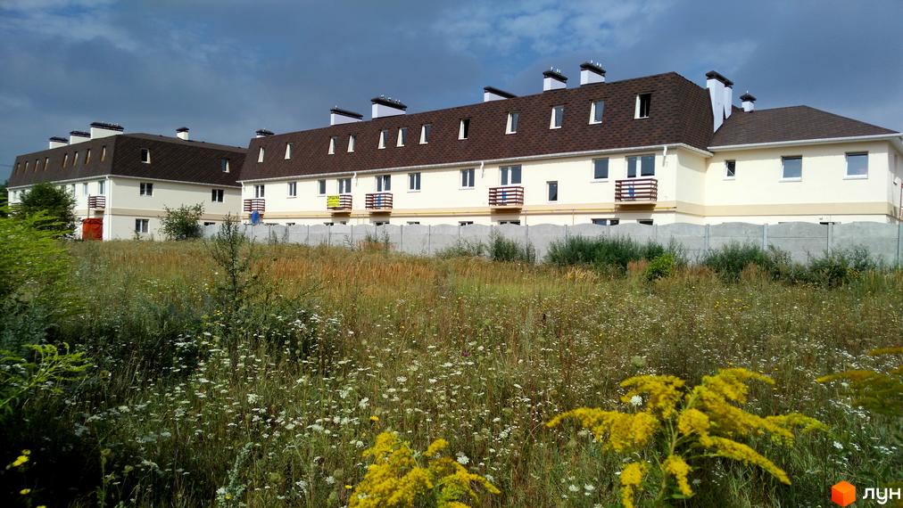 Зображення житлового комплексу з таунхаусами. Будинки з коричневими дахами та балконами, перед ними поле з рослинністю та бетонний паркан.