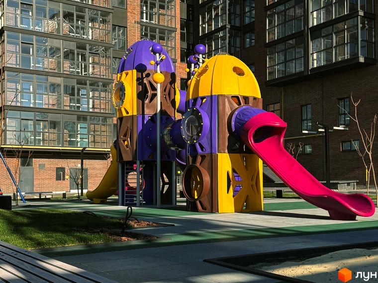 Житловий комплекс Стандарт: дитячий майданчик з яскравими гірками та будинок з цегляними та скляними фасадами. Прибудинкова територія з газоном та пісочницею.