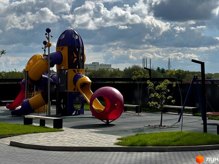Дитячий майданчик у житловому комплексі Стандарт з яскравими гірками та гойдалками. Навколо зелена трава та сучасне покриття.