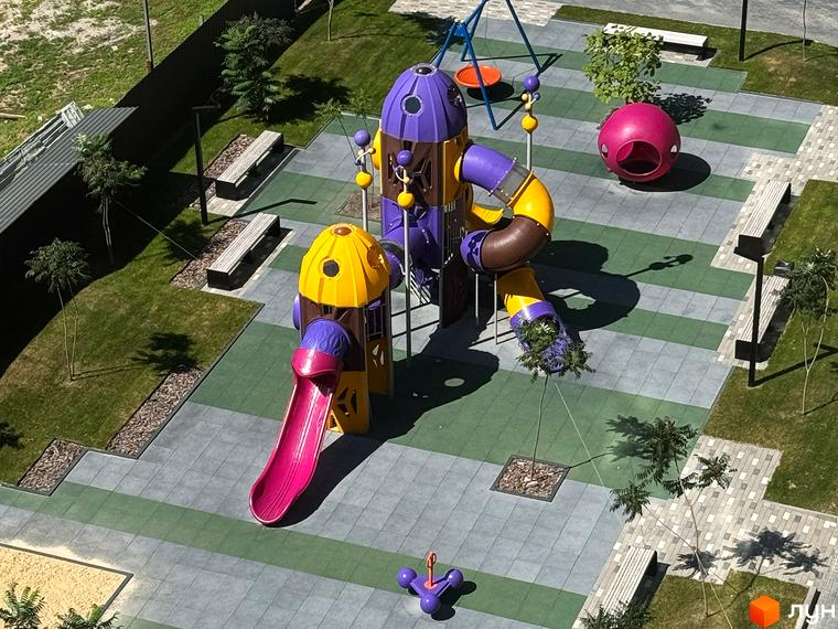 Дитячий майданчик у житловому комплексі Стандарт з яскравими гірками та гойдалками. Зелена прибудинкова територія з лавками та деревами.