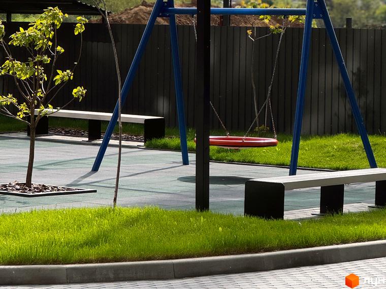 Дитячий майданчик з гойдалкою та лавками біля паркану в житловому комплексі Стандарт. Зелена трава та дерева навколо.