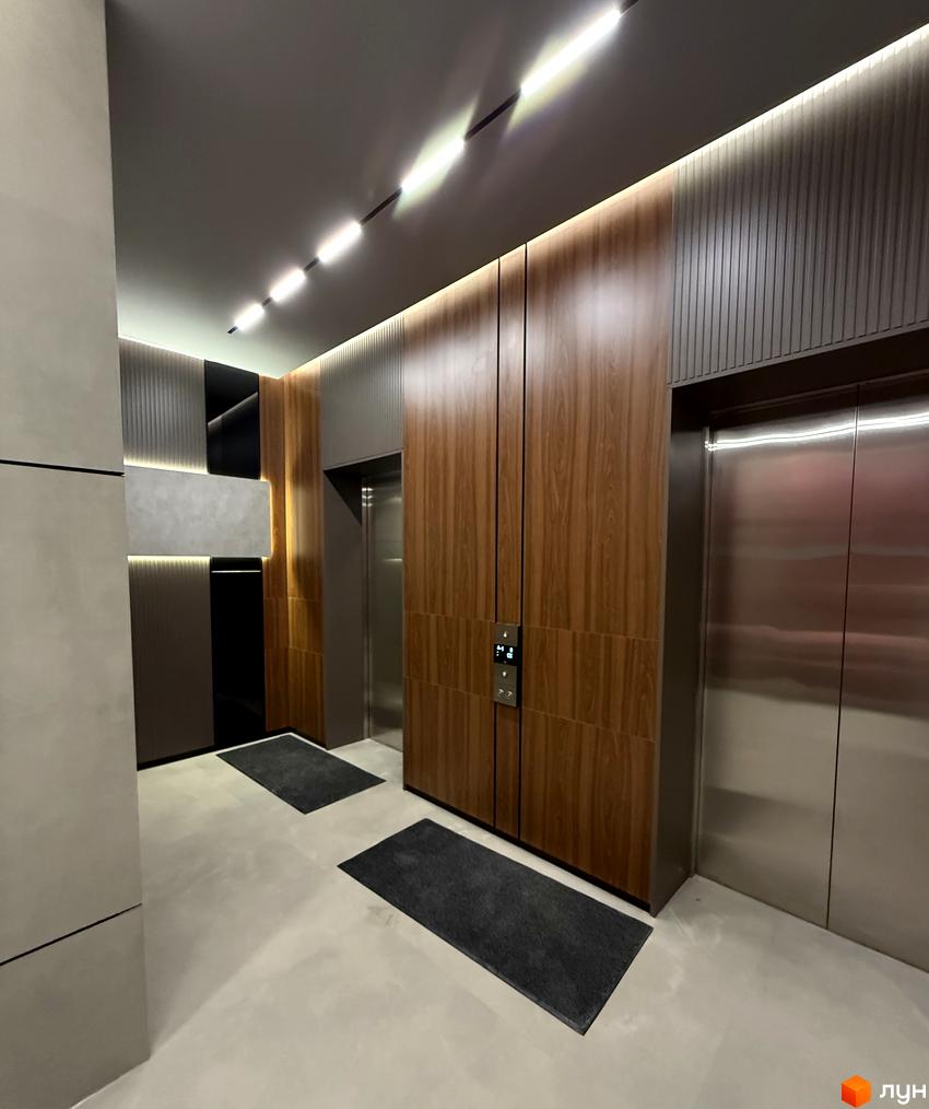 Інтер'єр холу новобудови ЖК Гімназист з двома ліфтами, оздоблений деревом та металом, з підсвічуванням стелі. Сучасний дизайн прибудинкової території.