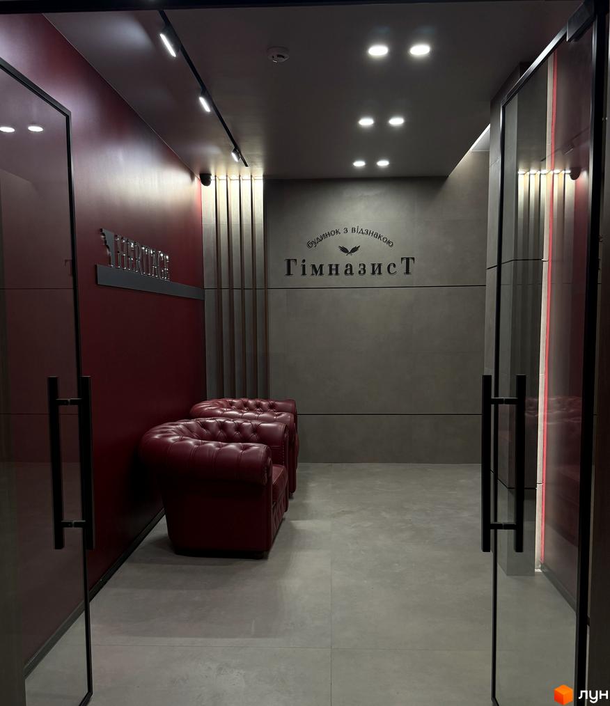 Житловий комплекс Гімназист: сучасний вхід з червоними диванами та логотипом. Просторий хол з сірими стінами та підлогою.