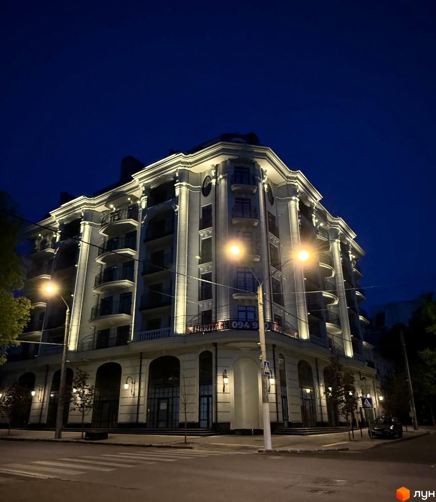 Новобудова ЖК Гімназист у вечірній час. Фасад будівлі з балконами та колонами, освітлений ліхтарями. Поруч дорога та пішохідний перехід.