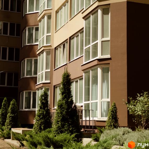 Будинок житлового комплексу Нова Європа з коричневими та бежевими фасадами, великими вікнами та зеленими ялинами на прибудинковій території.