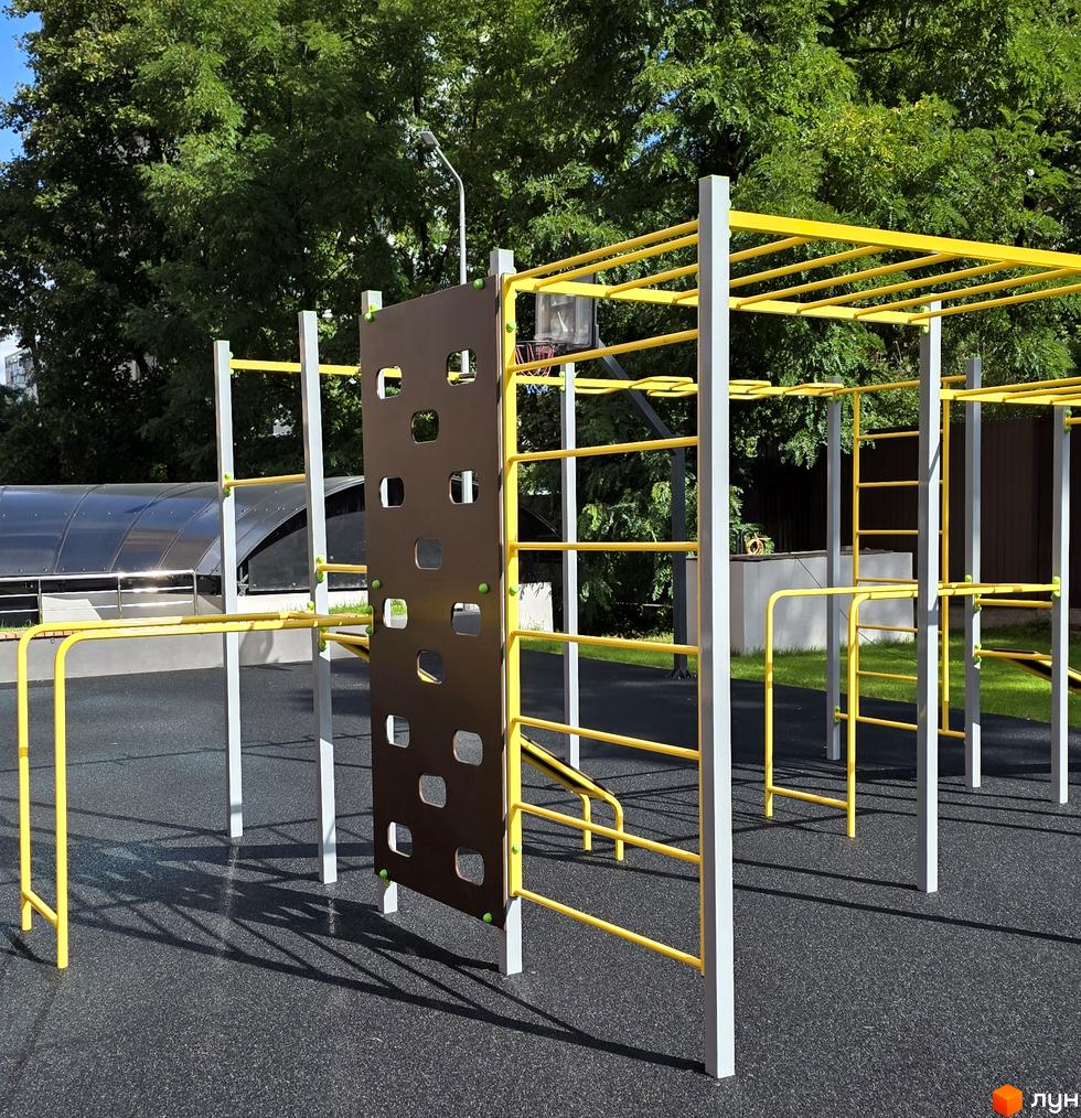 Дитячий майданчик з жовтими елементами та коричневою стіною для лазіння на фоні зелених дерев. Територія ЖК Eco Dream з безпечним покриттям.