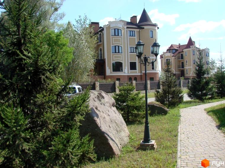 Зображення житлового комплексу. На фото видно будівлі з баштами та прибудинкову територію з доріжками та зеленими насадженнями.