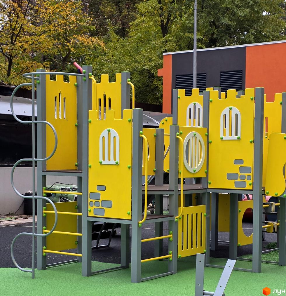 Дитячий майданчик житлового комплексу Eco Dream з яскравими жовтими елементами та сірою конструкцією, оточений зеленою травою та деревами.