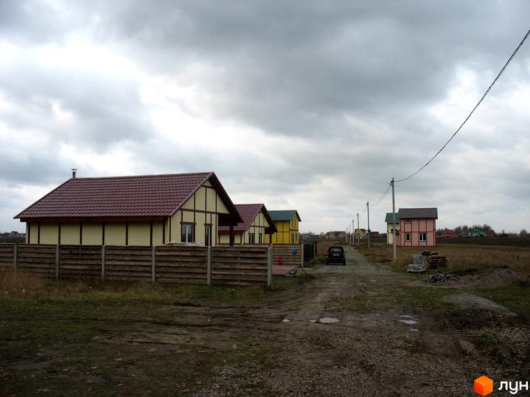 Житловий комплекс Під Горою з одноповерховими будинками з коричневими дахами та жовтими стінами. На передньому плані дерев'яний паркан, дорога та поле.