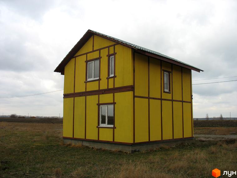 Жовтий двоповерховий будинок з коричневими смугами та вікнами. Будинок розташований на зеленій галявині, навколо видно поле та хмарне небо.