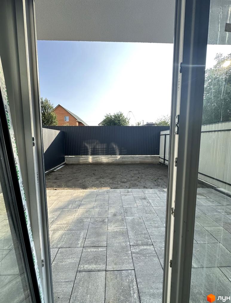 Вид на прибудинкову територію з вікна таунхаусу. На подвір'ї викладена плитка, за парканом знаходиться ділянка з землею.