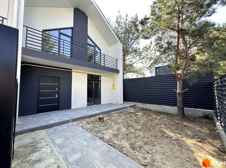 Білий будинок з чорними елементами, балконом та великими вікнами. Поруч доглянута прибудинкова територія з тротуаром та деревами.