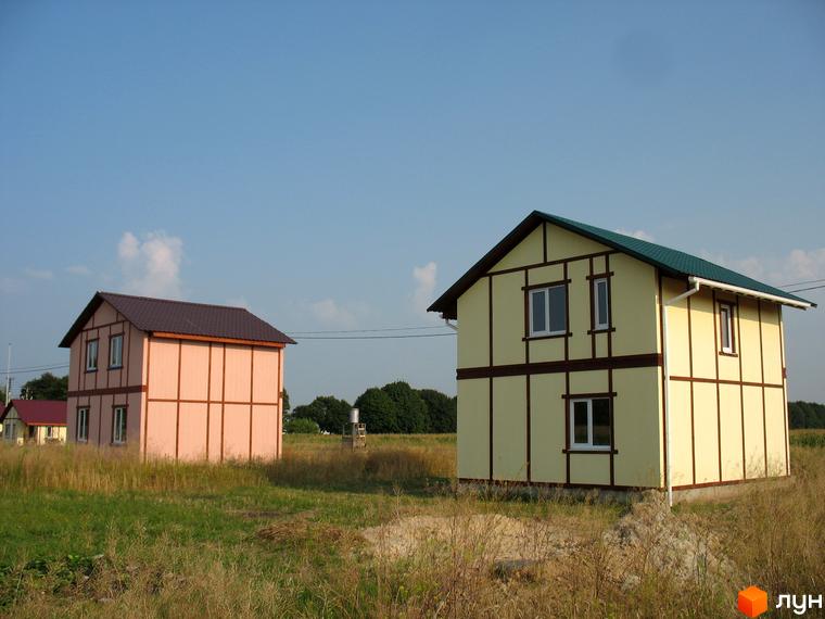 Двоповерховий будинок з жовтими стінами та коричневими декоративними балками. Поруч розташована ділянка з сухою травою та блакитне небо.