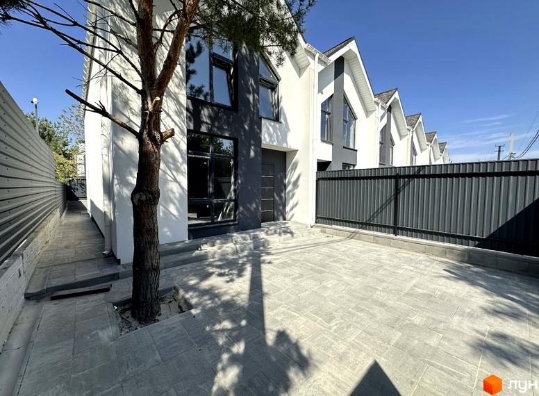 Фасад таунхаусу з великими вікнами та темними вставками. Біля будинку викладена бруківка, росте дерево та встановлено паркан.