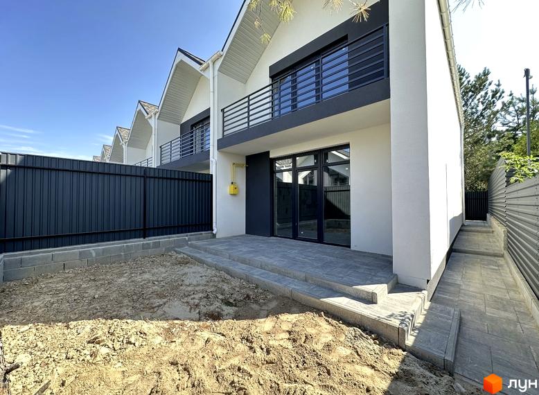 Білий таунхаус з чорними вікнами та балконом. Перед будинком розташована прибудинкова територія з тротуарною плиткою та огорожею.