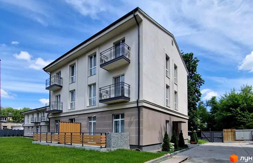 Зображення будинку ЖК Затишна Оселя з балконами та прилеглою територією з газоном та огорожею. На задньому плані видно блакитне небо з хмарами.