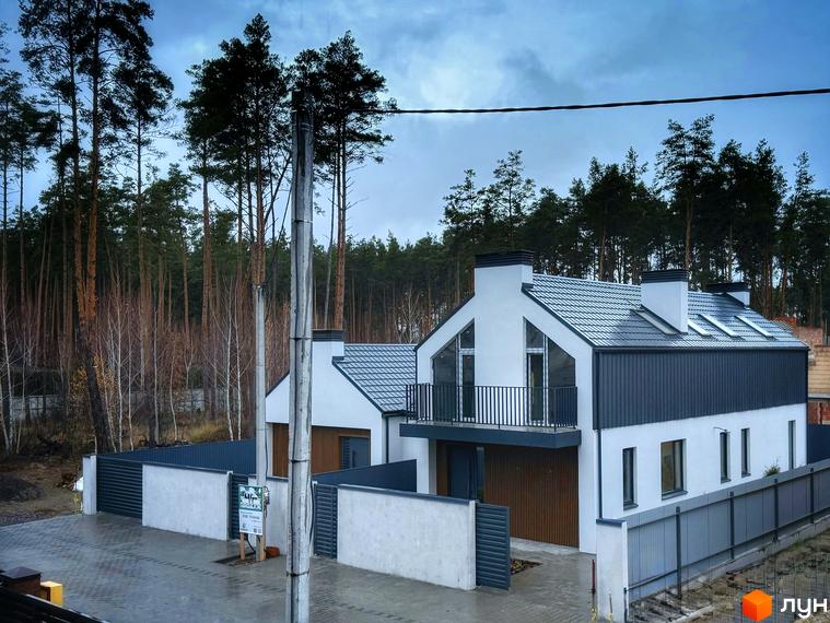 Двоповерховий будинок з білими стінами та темно-сірою покрівлею. На території будинку є паркан та дерева.