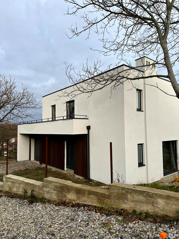 Білий будинок з плоским дахом, великими вікнами та дерев'яними панелями на фасаді. Перед будинком бетонний парапет та гравій.