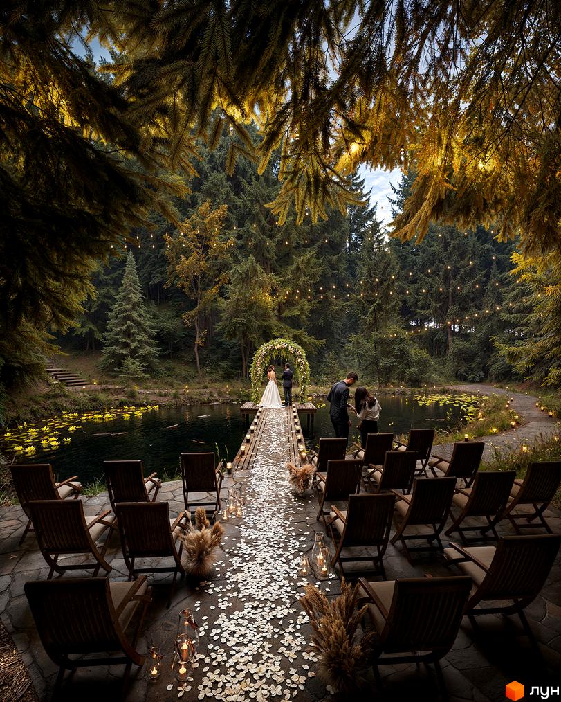 Весільна церемонія на дерев'яному пірсі над озером, прикрашена квітами та гірляндами. На березі розташовані дерев'яні стільці для гостей.